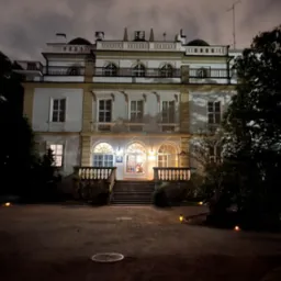 Elewacja dwupiętrowego budynku z balkonem i schodami wejściowymi, oświetlona nocą. Widoczne lampy ogrodowe i drzewa. Budynek w stylu klasycystycznym, z elementami dekoracyjnymi.