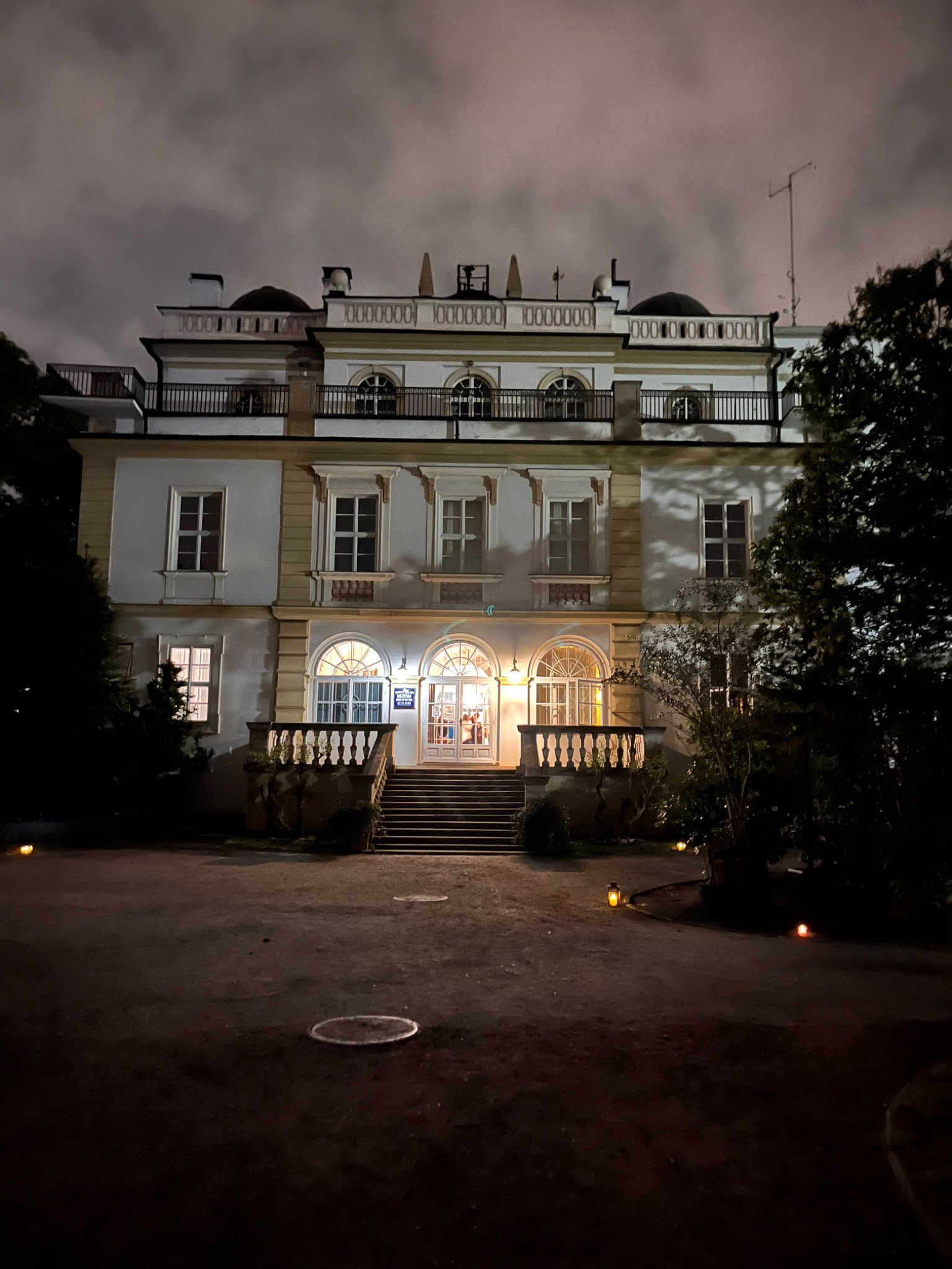 Elewacja dwupiętrowego budynku z balkonem i schodami wejściowymi, oświetlona nocą. Widoczne lampy ogrodowe i drzewa. Budynek w stylu klasycystycznym, z elementami dekoracyjnymi.