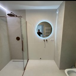 FLIZBUD SPÓŁKA Z OGRANICZONĄ ODPOWIEDZIALNOŚCIĄ - Nowoczesna łazienka z kabiną prysznicową typu walk-in, okrągłym lustrem z podświetleniem LED i armaturą w kolorze miedzi. Częściowo wykończone ściany.