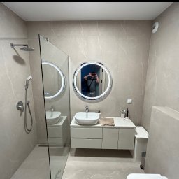 FLIZBUD SPÓŁKA Z OGRANICZONĄ ODPOWIEDZIALNOŚCIĄ - Nowoczesna łazienka w beżowych płytkach z kabiną prysznicową typu walk-in, okrągłym lustrem z podświetleniem LED i białą umywalką nablatową.