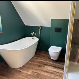 FLIZBUD SPÓŁKA Z OGRANICZONĄ ODPOWIEDZIALNOŚCIĄ - Nowoczesna łazienka na poddaszu: wolnostojąca wanna, toaleta i kabina prysznicowa z złotymi detalami na tle butelkowej zieleni ścian i drewnopodobnej podłogi.