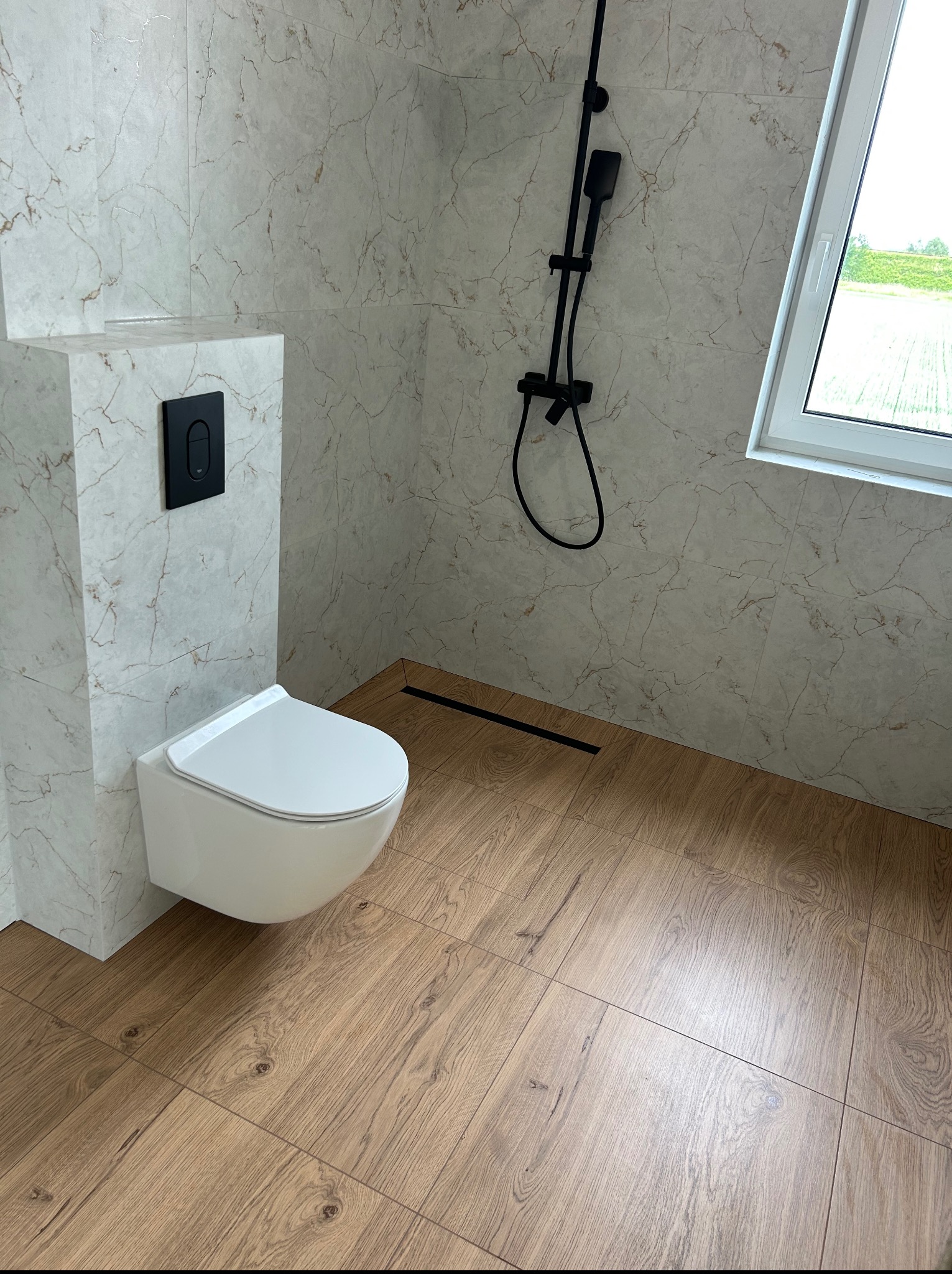 Nowoczesna łazienka z beżowymi płytkami imitującymi marmur, drewnopodobną podłogą i czarną armaturą. Uwagę zwraca podwieszana miska WC i okno z widokiem na zieleń.