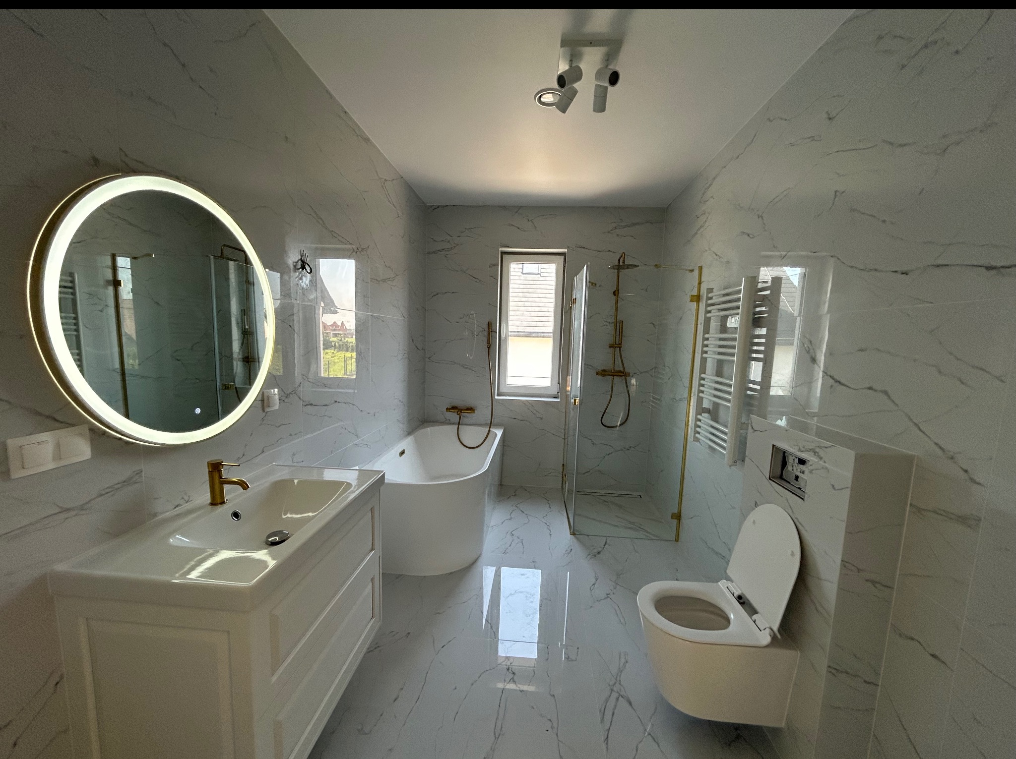 Nowoczesna łazienka z marmurowymi płytkami, okrągłym lustrem z podświetleniem LED, białą armaturą i złotymi akcentami, widok na okno i kabinę prysznicową.