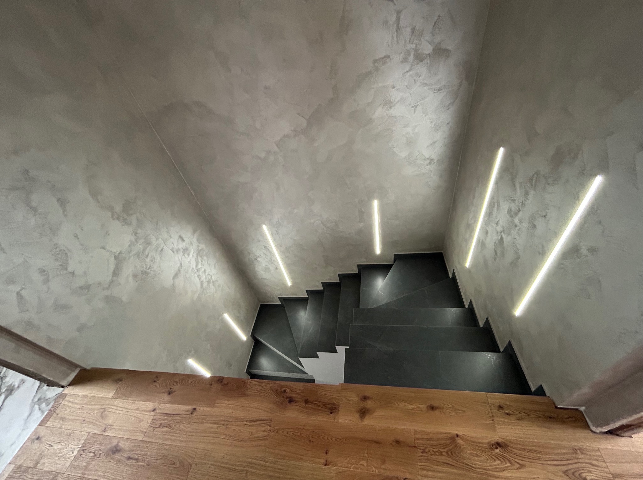 Nowoczesne schody z szarymi stopniami i oświetleniem LED na ścianach z efektem dekoracyjnym. Drewniana podłoga na dole. Ujęcie z góry.