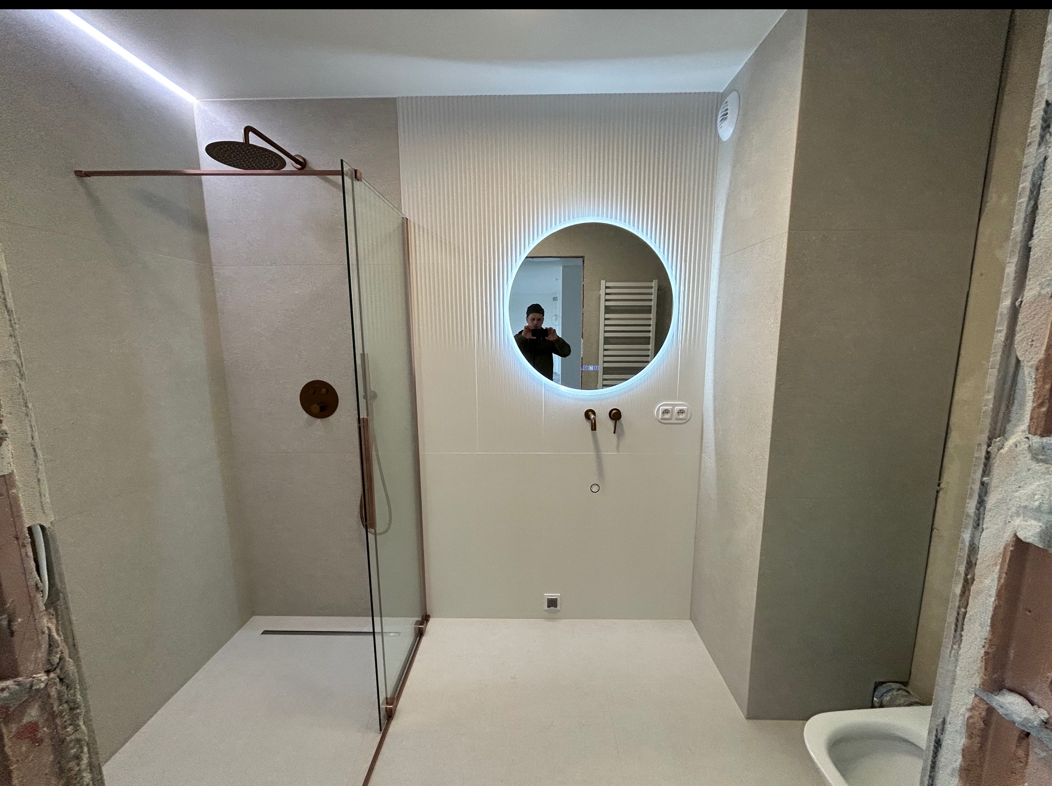 Nowoczesna łazienka z kabiną prysznicową typu walk-in, okrągłym lustrem z podświetleniem LED i armaturą w kolorze miedzi. Częściowo wykończone ściany.