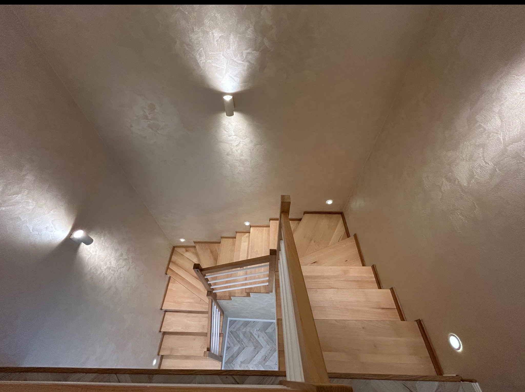 Klatka schodowa z drewnianymi schodami, widok z góry. Ściany wykończone dekoracyjnym tynkiem w odcieniach beżu, oświetlone punktowymi lampami.