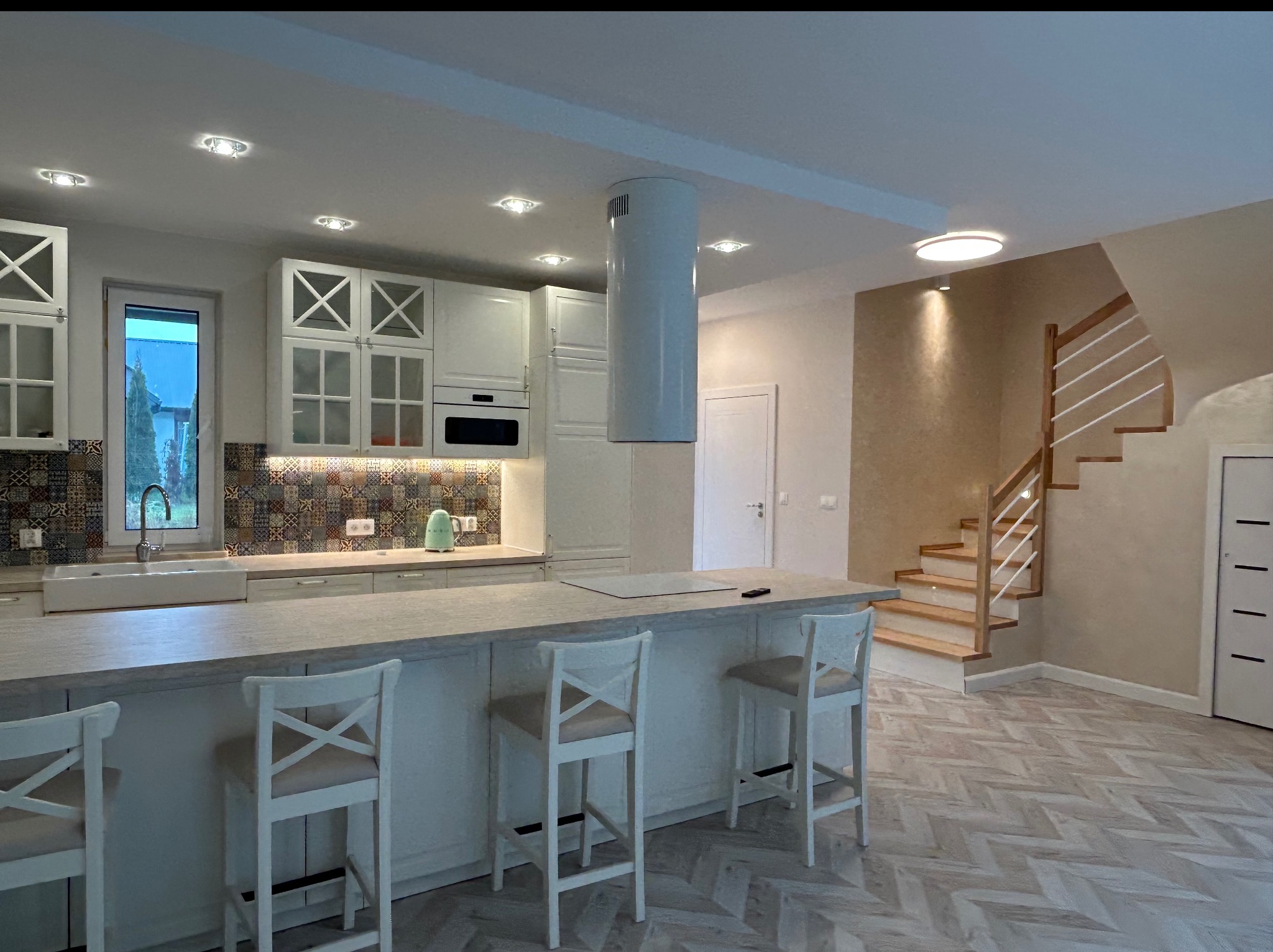 Jasna, przestronna kuchnia z wyspą i schodami w tle, Kraków. Widoczne białe meble, wzorzyste płytki nad blatem i parkiet w jodełkę.