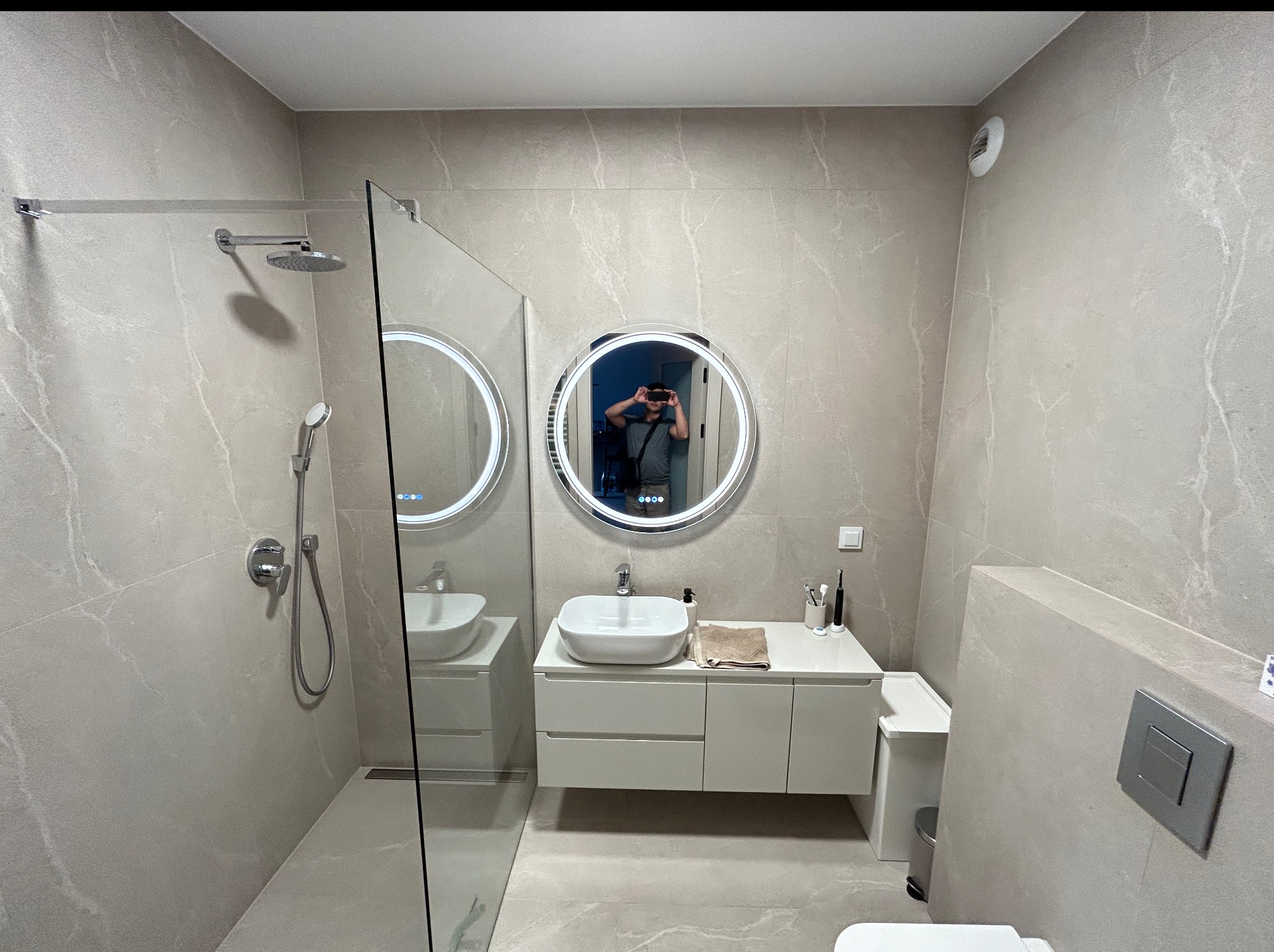 Nowoczesna łazienka w beżowych płytkach z kabiną prysznicową typu walk-in, okrągłym lustrem z podświetleniem LED i białą umywalką nablatową.