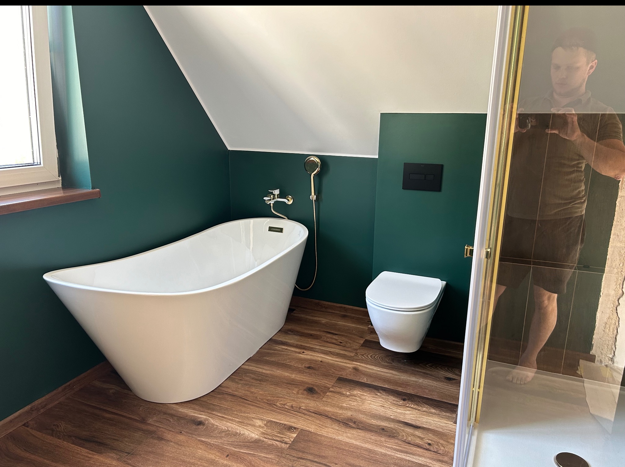 Nowoczesna łazienka na poddaszu: wolnostojąca wanna, toaleta i kabina prysznicowa z złotymi detalami na tle butelkowej zieleni ścian i drewnopodobnej podłogi.