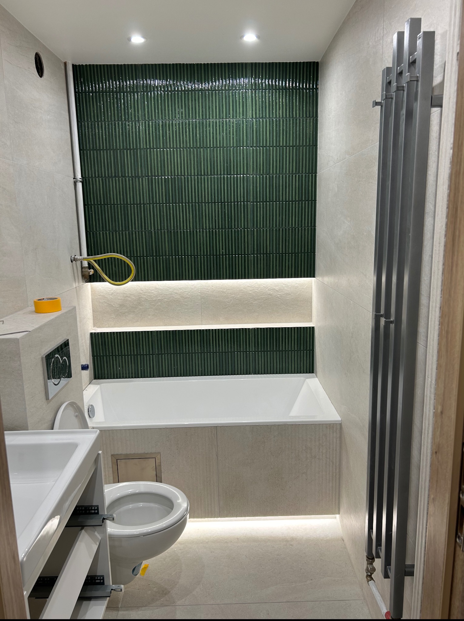 Nowoczesna łazienka z zieloną, pionową mozaiką nad wanną, podświetleniem LED i grzejnikiem drabinkowym. Umywalka z szafką i toaleta w neutralnej kolorystyce.