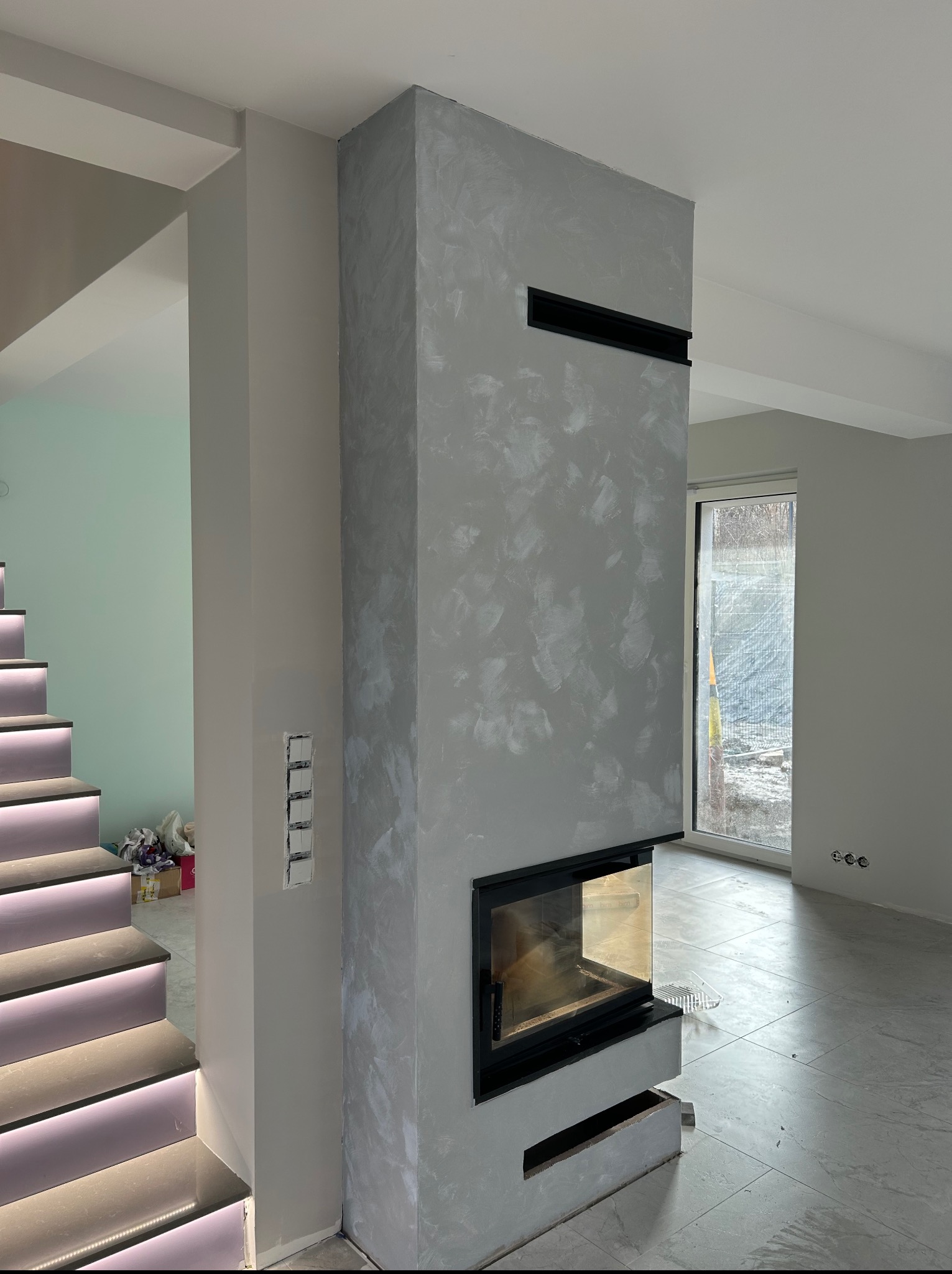 Nowoczesny kominek z szarą, dekoracyjną strukturą w jasnym wnętrzu. Widoczne schody z ledowym podświetleniem i okno. Minimalistyczny design.