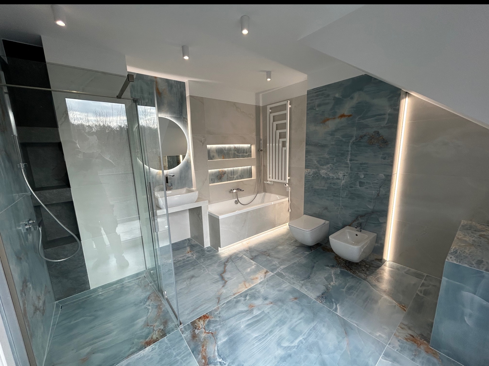 Nowoczesna łazienka na poddaszu z niebieską armaturą i oświetleniem LED. Kabina prysznicowa ze szklaną ścianą, umywalka, wanna, bidet i toaleta. Wykończenie w stylu marmuru.