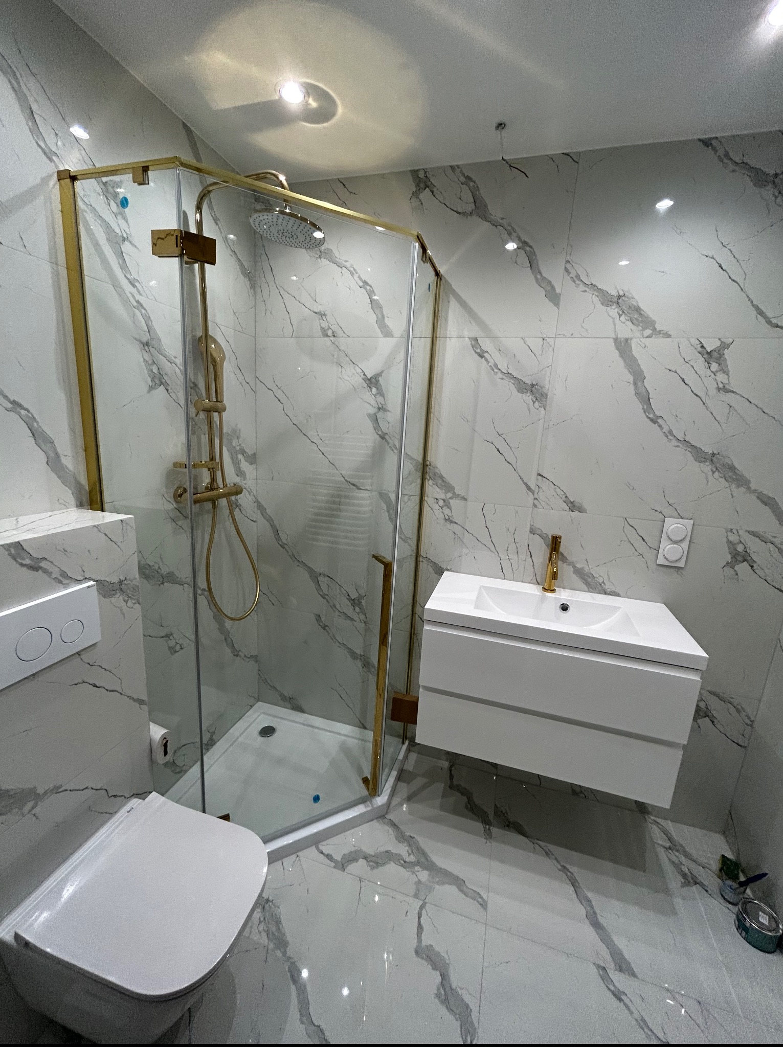 Nowoczesna łazienka z kabiną prysznicową w złotej ramie, białą umywalką i toaletą. Marmurowe płytki na ścianach i podłodze. Minimalistyczny design, punktowe oświetlenie.