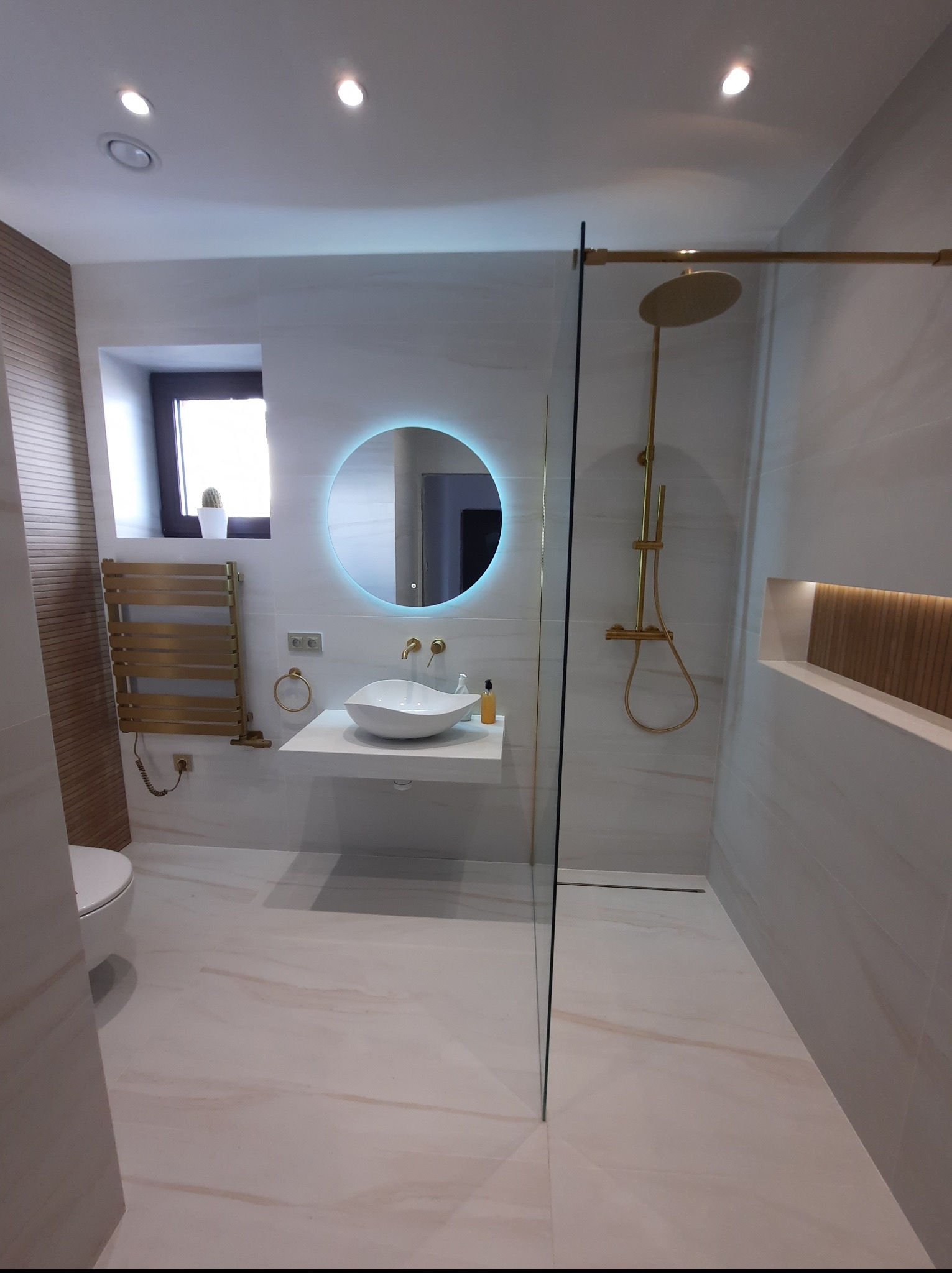 Nowoczesna łazienka z białymi płytkami imitującymi marmur, okrągłym lustrem z podświetleniem LED i złotą armaturą, prezentująca minimalistyczny design.