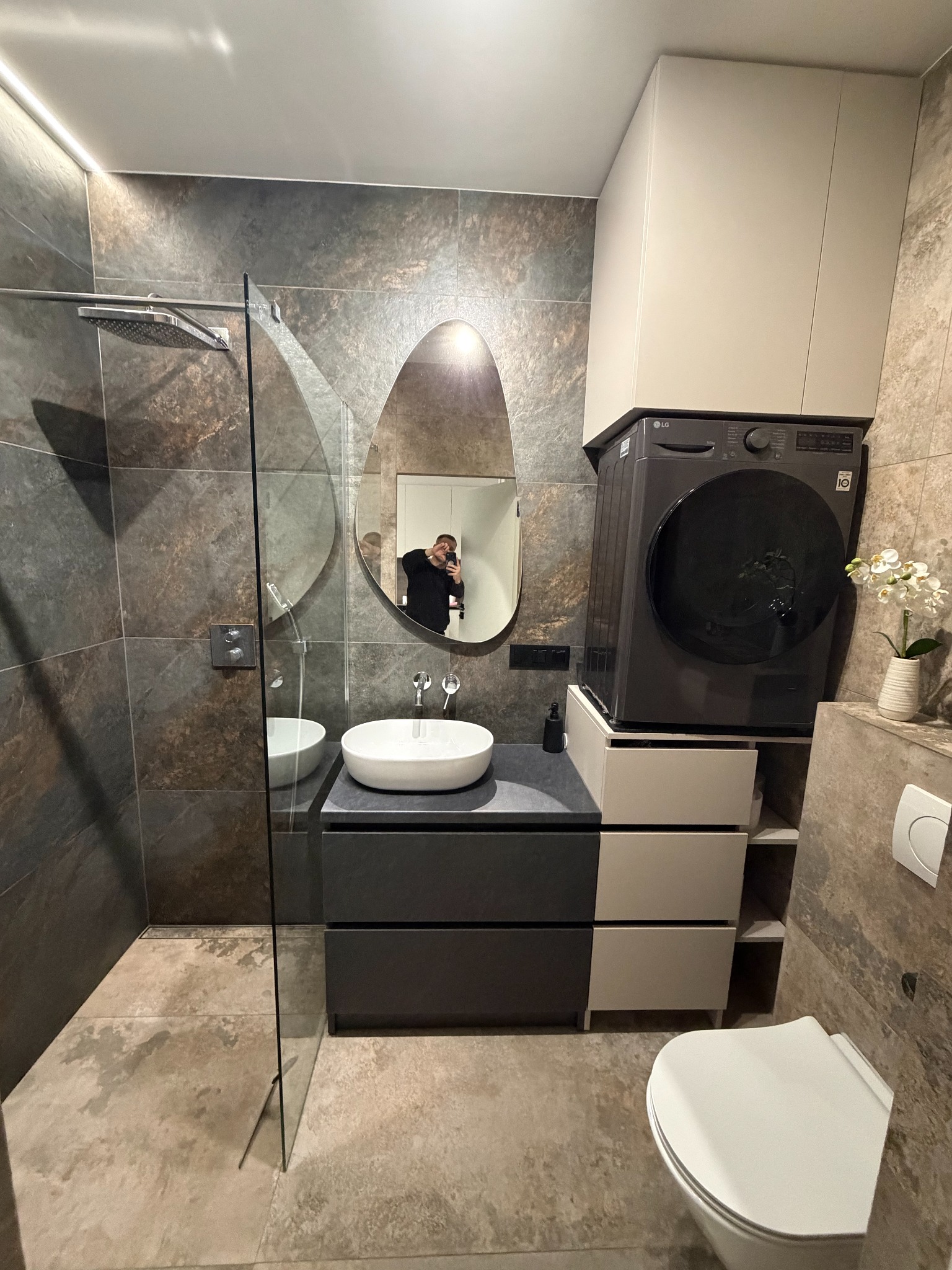 Nowoczesna łazienka z kabiną prysznicową typu walk-in, owalnym lustrem i pralką LG umieszczoną nad szafką z szufladami. Ciemne płytki imitujące kamień.