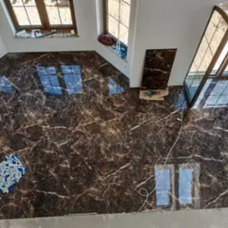 Układanie płytek podłogowych imitujących marmur w pomieszczeniu z oknami. Widoczne narzędzia i nieułożone płytki, co wskazuje na etap prac wykończeniowych.