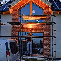 Arko - Murowanie elewacji domu z cegły klinkierowej. Widoczne rusztowanie i pracownik na tle budynku z drewnianą stolarką okienną.