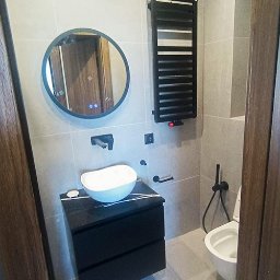 Arko - Nowoczesna łazienka z okrągłym lustrem, umywalką na czarnej szafce i grzejnikiem drabinkowym. Szare płytki i czarne akcenty tworzą elegancki wystrój.