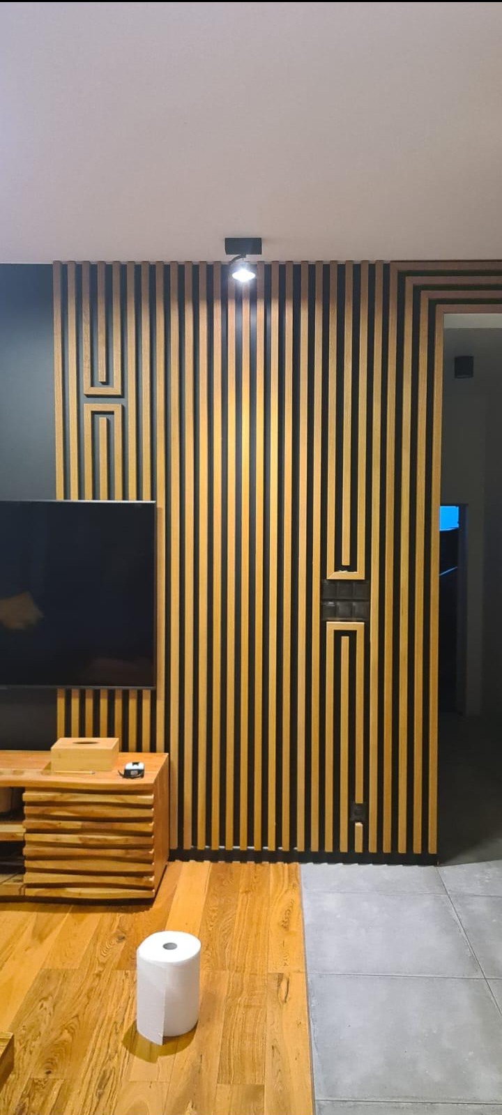 Nowoczesna ściana z drewnianych lameli z ukrytymi drzwiami, podświetlana lampą punktową. Element dekoracyjny zintegrowany z przejściem do innego pomieszczenia.