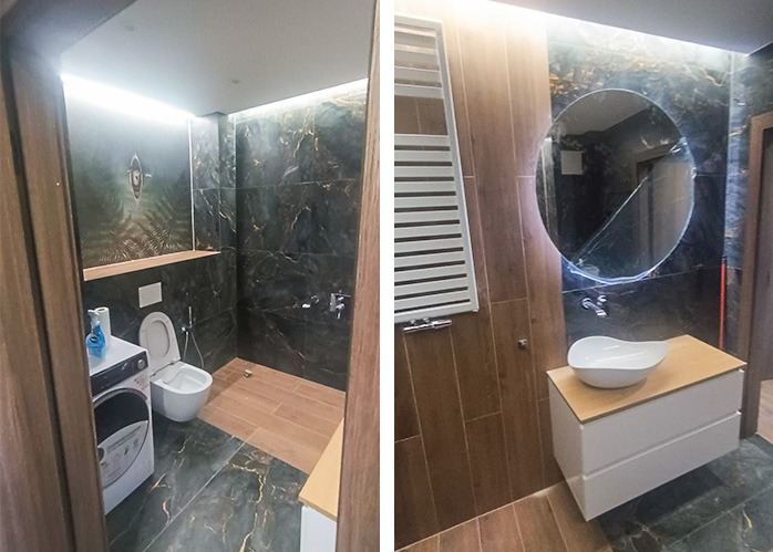 Nowoczesna łazienka z czarnymi płytkami imitującymi marmur, drewnianymi akcentami i okrągłym lustrem z podświetleniem LED. Widoczna pralka i grzejnik.