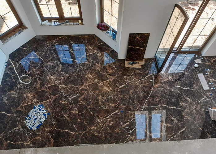 Układanie płytek podłogowych imitujących marmur w pomieszczeniu z oknami. Widoczne narzędzia i nieułożone płytki, co wskazuje na etap prac wykończeniowych.