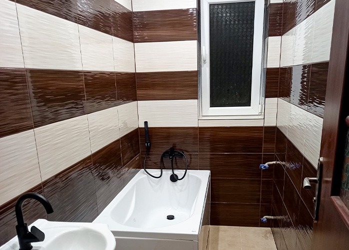 Wykończona łazienka z brązowo-białymi płytkami w poziome pasy, czarną armaturą i białym oknem. Widoczny fragment umywalki i drzwi.