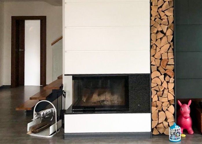 Nowoczesny kominek z białymi panelami i czarnym obramowaniem, obok ściana z ułożonym drewnem. Widoczne schody i drzwi w tle. Dekoracje na podłodze.
