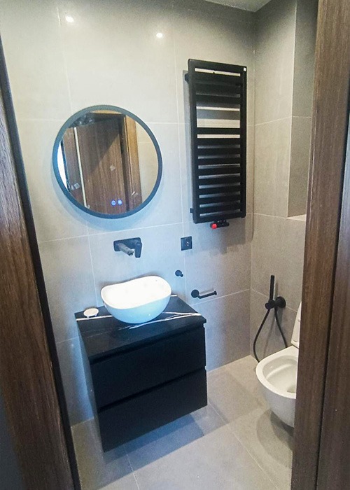 Nowoczesna łazienka z okrągłym lustrem, umywalką na czarnej szafce i grzejnikiem drabinkowym. Szare płytki i czarne akcenty tworzą elegancki wystrój.