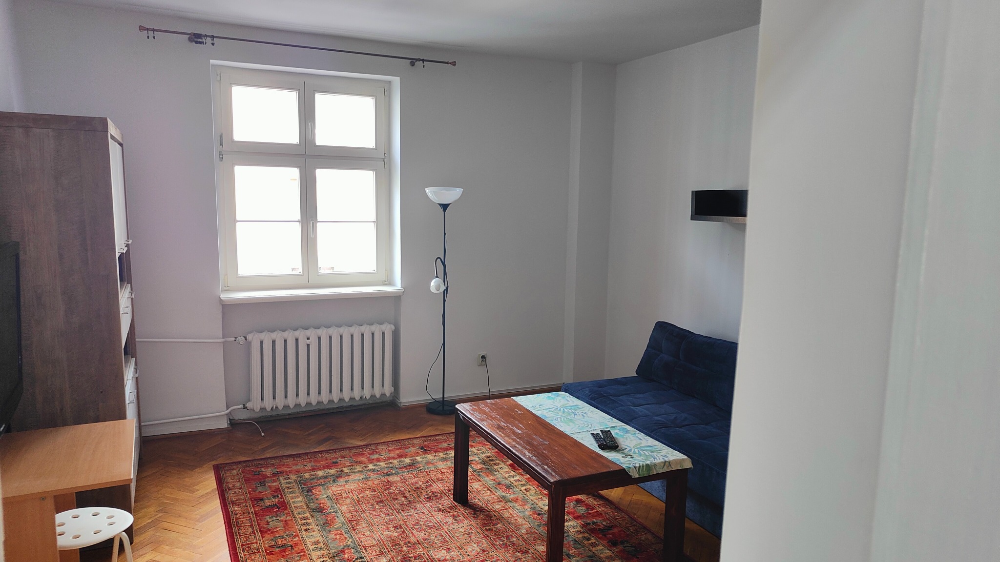 Jasne wnętrze pokoju z drewnianą podłogą i dywanem. Widoczne okno, grzejnik, szafa, lampa stojąca i niebieska sofa. Na stole leżą piloty.