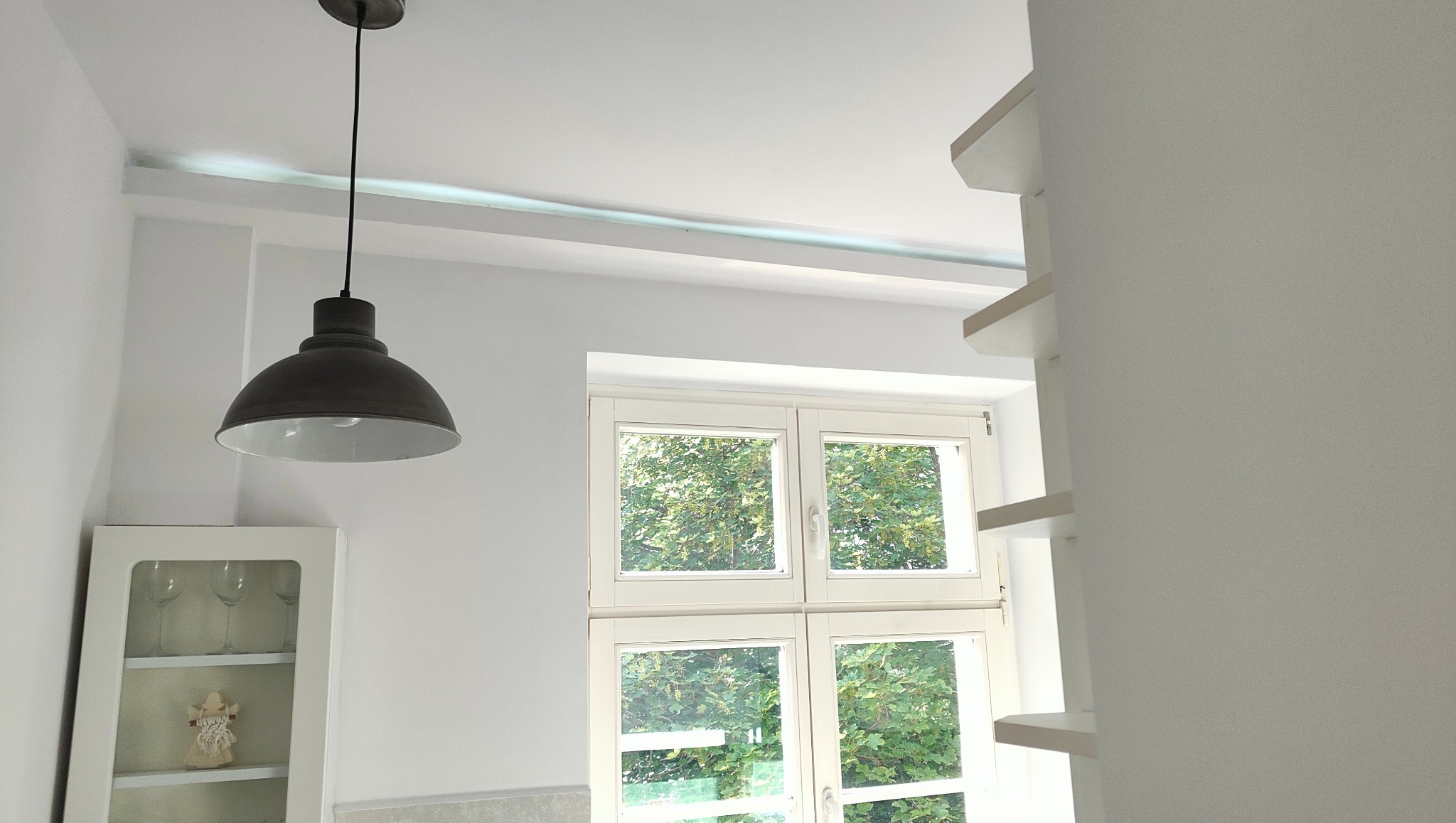 Jasne wnętrze po remoncie: białe ściany, okno z widokiem na zieleń, lampa w stylu loft i biały regał. Minimalistyczny design, czystość i świeżość.