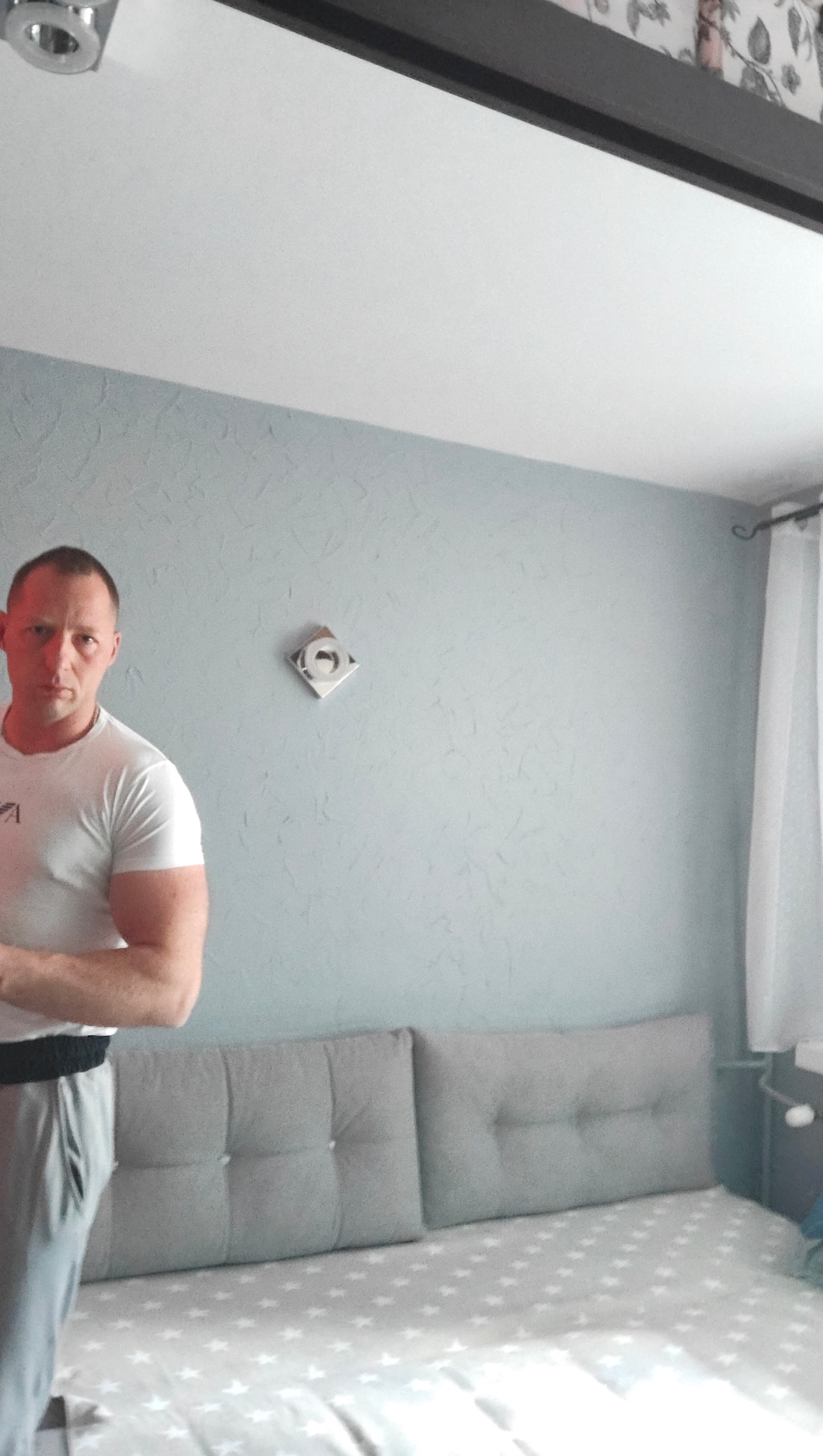 Ściana po malowaniu z fakturą, szara sofa z guzikami, lampa kwadratowa. Mężczyzna w białej koszulce stoi z lewej strony kadru. Nowoczesne wykończenie wnętrz.