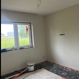 WykończTo - Pokój w trakcie wykańczania: szpachlowane ściany, przygotowana podłoga z widoczną instalacją grzewczą, okno z narzędziami, wiadro farby, poziomica laserowa.