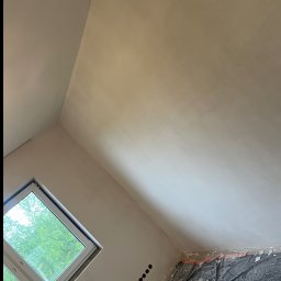 WykończTo - Ściana i sufit po szpachlowaniu, widoczne gniazdka elektryczne i okno z widokiem na zieleń. Podłoga zabezpieczona folią ochronną. Ukończony etap przygotowania do malowania.