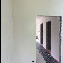 WykończTo - Wnętrze po szpachlowaniu: gładkie, białe ściany, przygotowane do malowania. Widoczne otwory drzwiowe i folia ochronna na podłodze. Minimalistyczny kadr, surowy stan wykończenia.