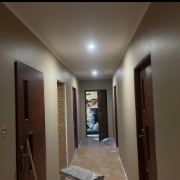 WykończTo - Korytarz po malowaniu ścian na beżowo, widoczne drzwi i sufit z oświetleniem. Na podłodze drabina malarska. Widok w głąb pomieszczenia.