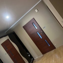 WykończTo - Korytarz po remoncie z dwiema ciemnymi, drewnianymi drzwiami z przeszkleniami. Ściany w odcieniach beżu, jasna podłoga. Widoczne ślady po pracach wykończeniowych.