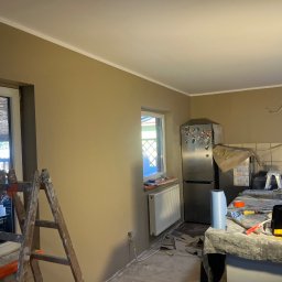 WykończTo - Kuchnia po malowaniu ścian na beżowo z białą listwą sufitową. Widoczne okno, drabina malarska i zabezpieczone meble. Ślady prac remontowych na podłodze.