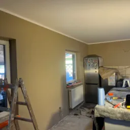 Kuchnia po malowaniu ścian na beżowo z białą listwą sufitową. Widoczne okno, drabina malarska i zabezpieczone meble. Ślady prac remontowych na podłodze.