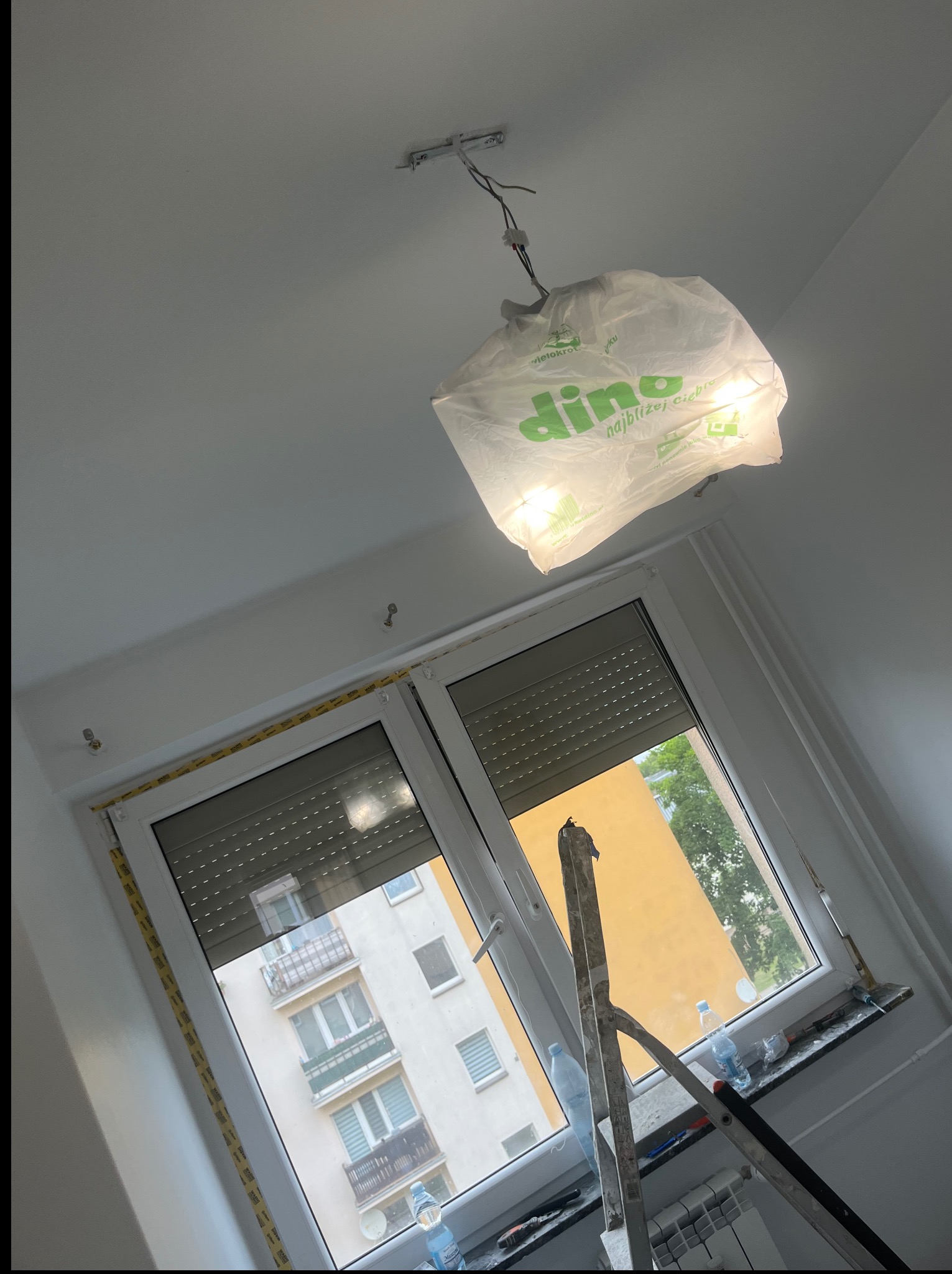 Białe wnętrze w trakcie malowania: zabezpieczona lampa pod sufitem, okno z roletami, drabina oparta o parapet, widoczne ślady prac malarskich.
