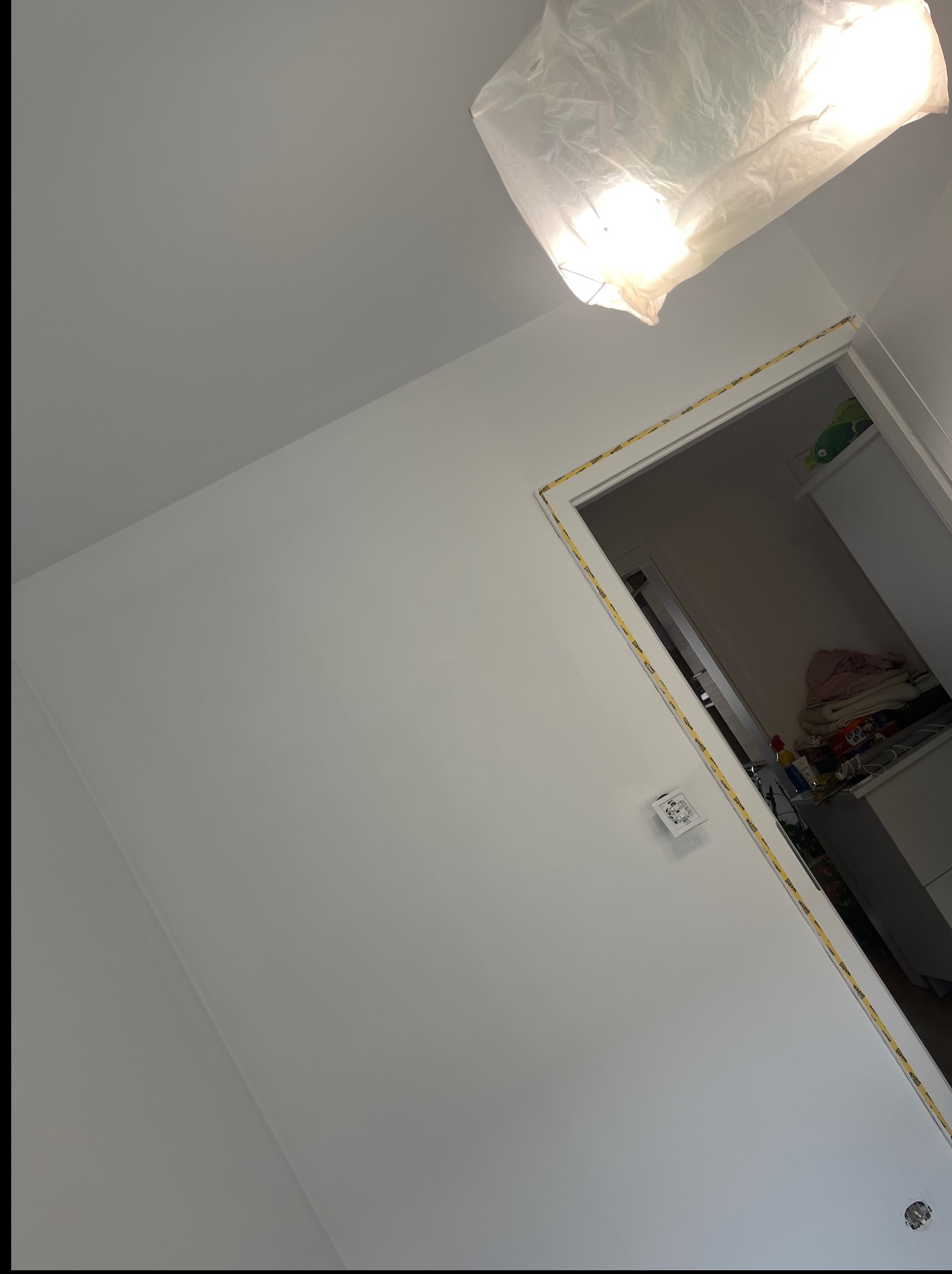 Świeżo pomalowane, białe ściany pokoju z zabezpieczonymi taśmą framugami drzwiowymi i zakrytym źródłem światła u góry kadru. Widoczny fragment gniazdka elektrycznego.