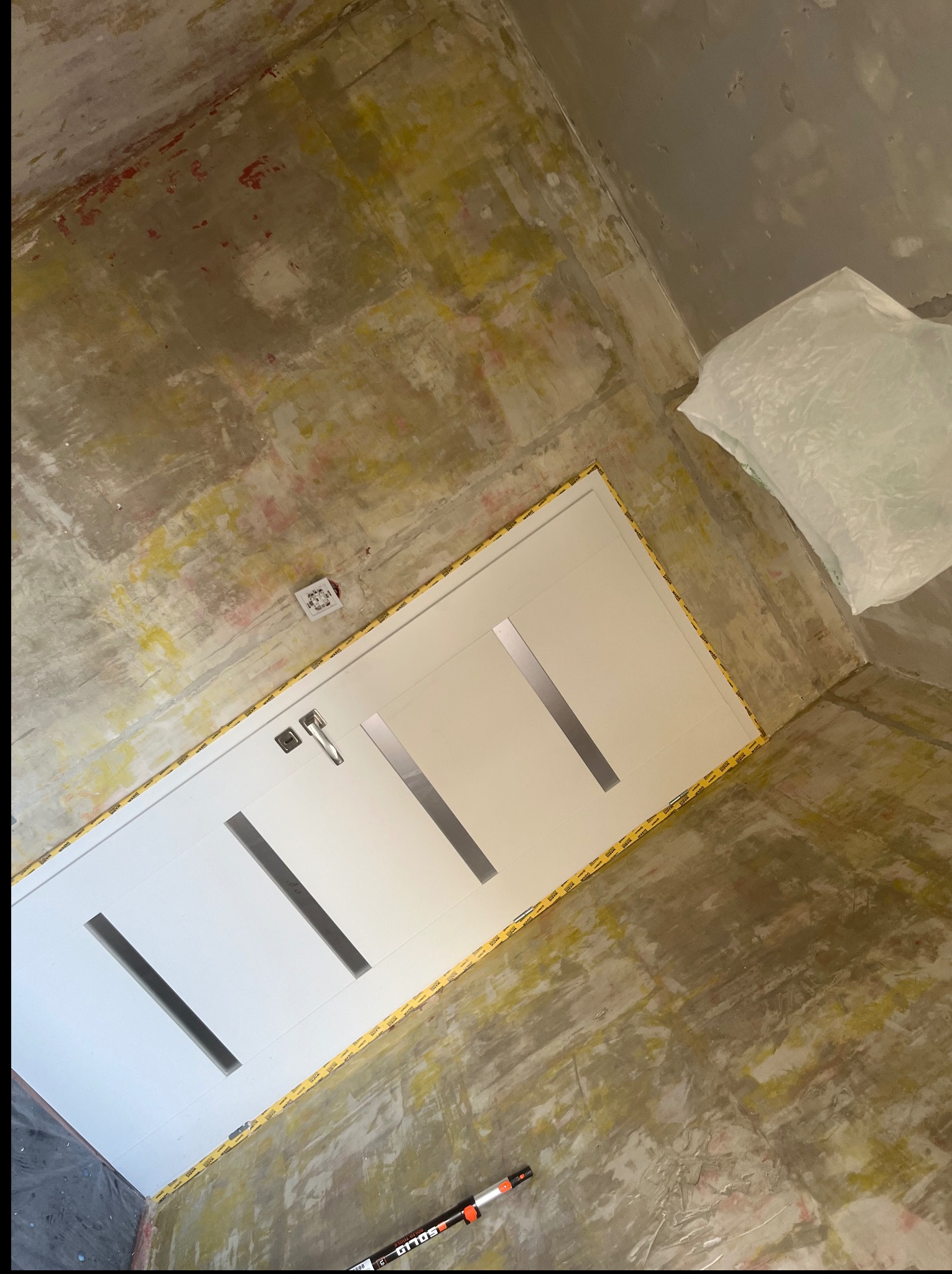 Białe drzwi z czarnymi wstawkami, zabezpieczone taśmą, w surowym wnętrzu w trakcie remontu. Widoczny fragment podłogi i ściany z widocznymi śladami prac wykończeniowych.