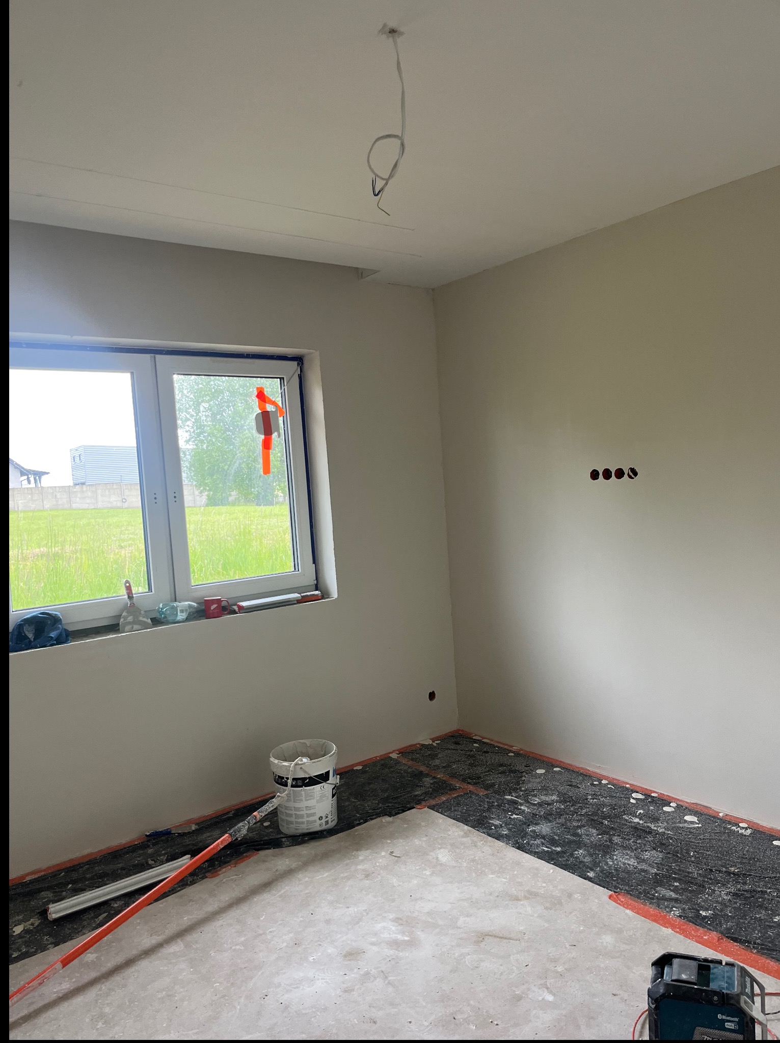 Pokój w trakcie wykańczania: szpachlowane ściany, przygotowana podłoga z widoczną instalacją grzewczą, okno z narzędziami, wiadro farby, poziomica laserowa.