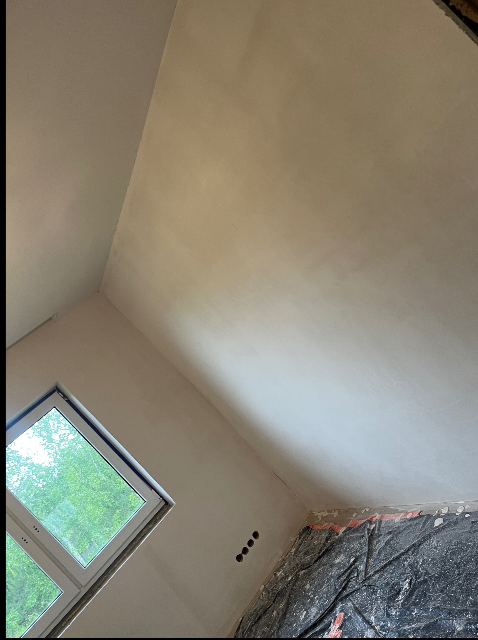 Ściana i sufit po szpachlowaniu, widoczne gniazdka elektryczne i okno z widokiem na zieleń. Podłoga zabezpieczona folią ochronną. Ukończony etap przygotowania do malowania.