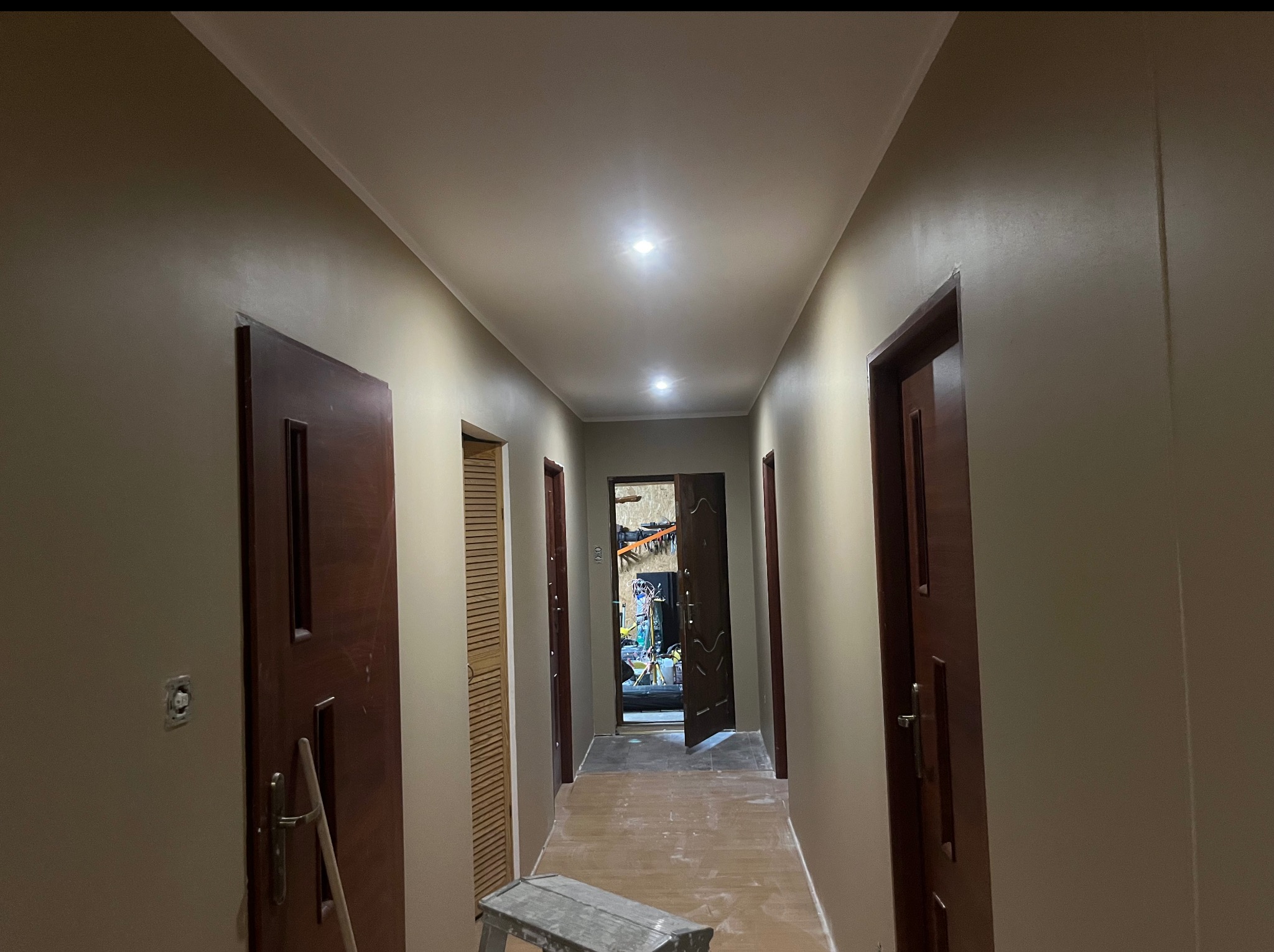 Korytarz po malowaniu ścian na beżowo, widoczne drzwi i sufit z oświetleniem. Na podłodze drabina malarska. Widok w głąb pomieszczenia.