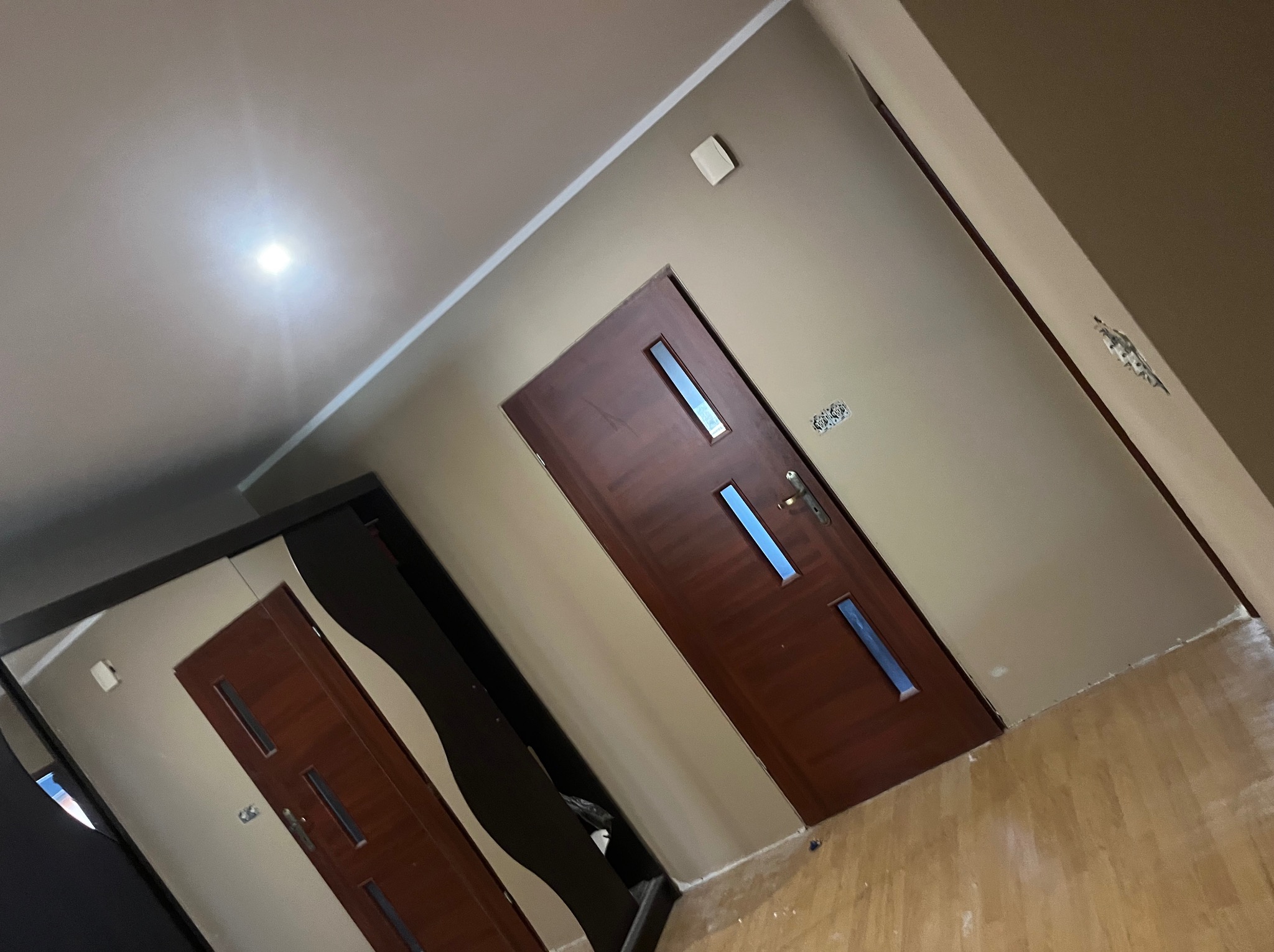 Korytarz po remoncie z dwiema ciemnymi, drewnianymi drzwiami z przeszkleniami. Ściany w odcieniach beżu, jasna podłoga. Widoczne ślady po pracach wykończeniowych.