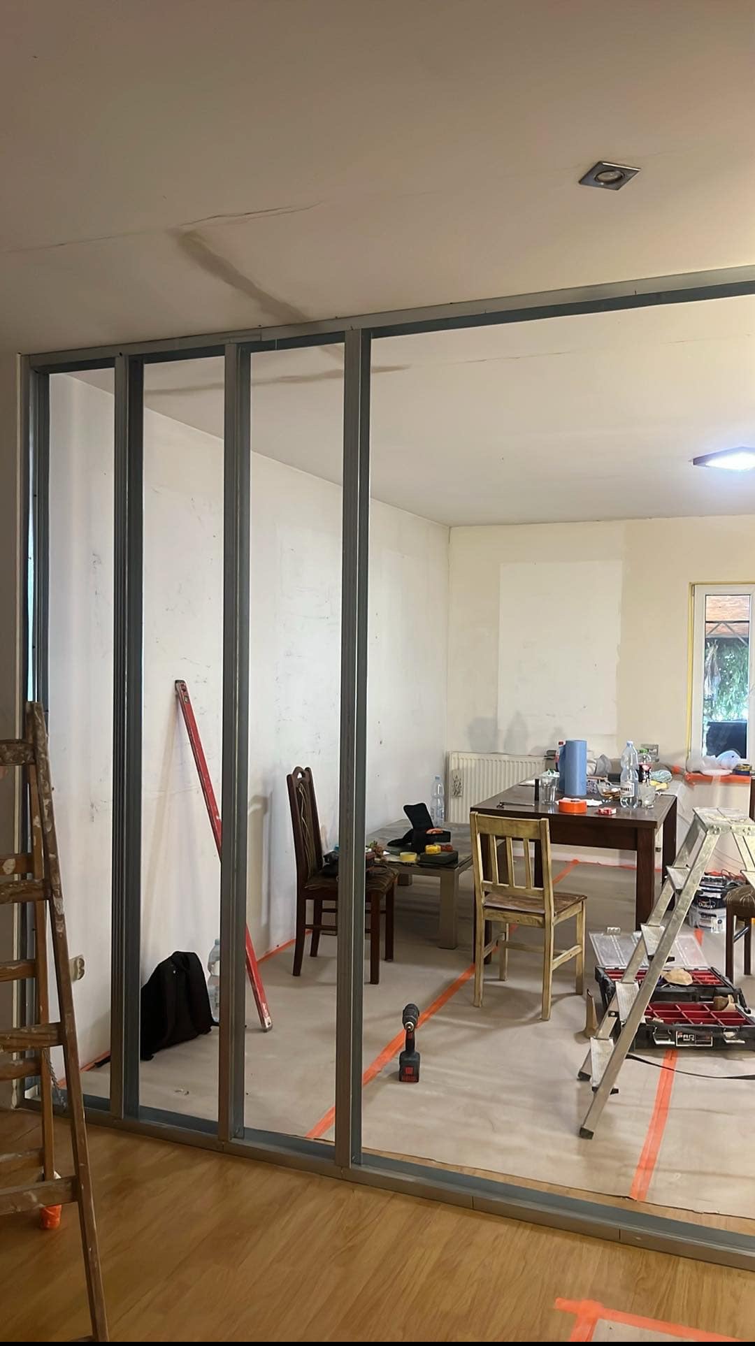 Metalowa konstrukcja ścianki działowej w trakcie montażu w pomieszczeniu. Widoczne narzędzia i drabina. Podłoga zabezpieczona folią. Widok na wnętrze pokoju.