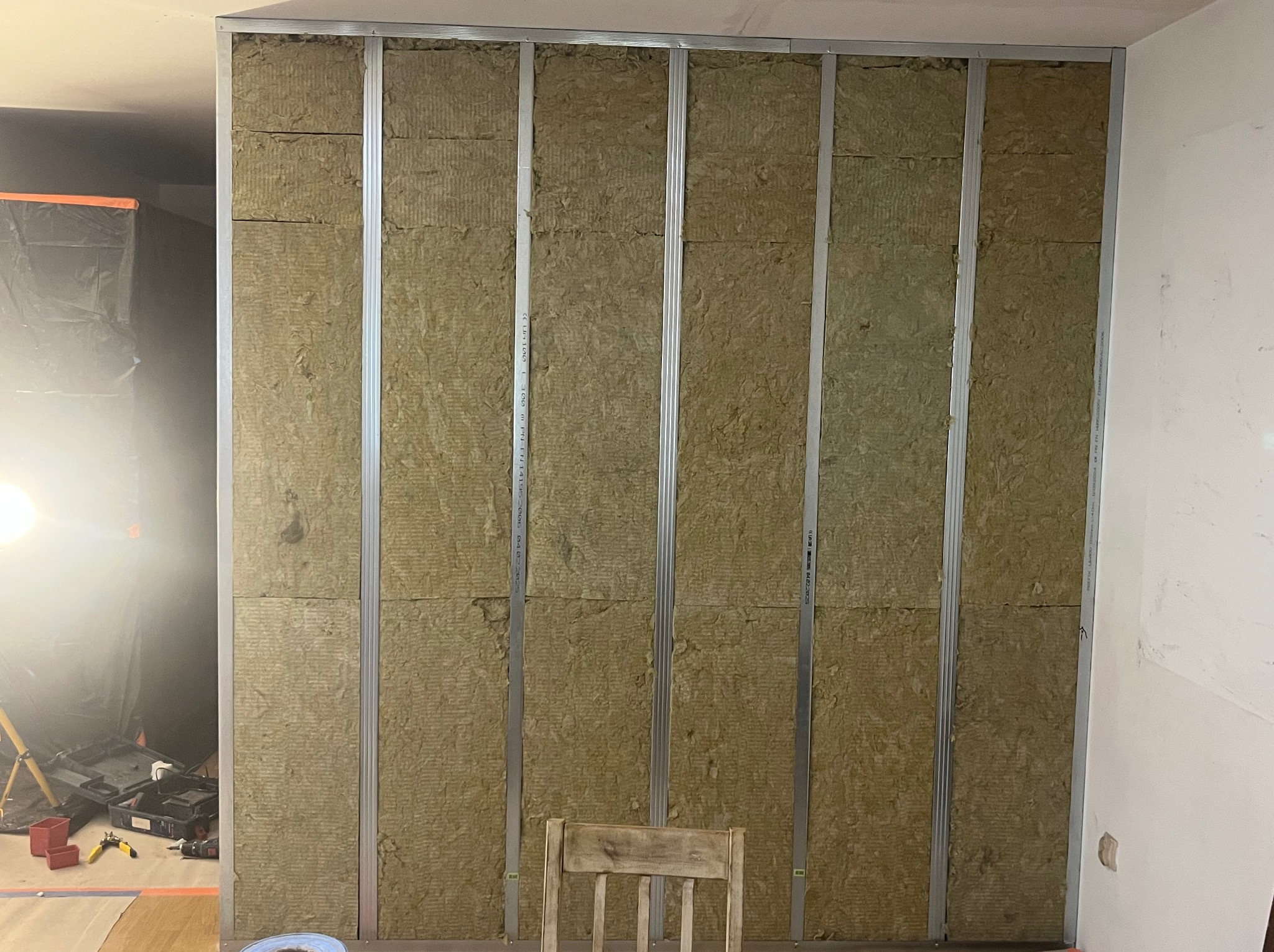 Montaż izolacji z wełny mineralnej w metalowym stelażu ściany działowej. Widoczne narzędzia i krzesło. Oświetlenie budowlane po lewej stronie.