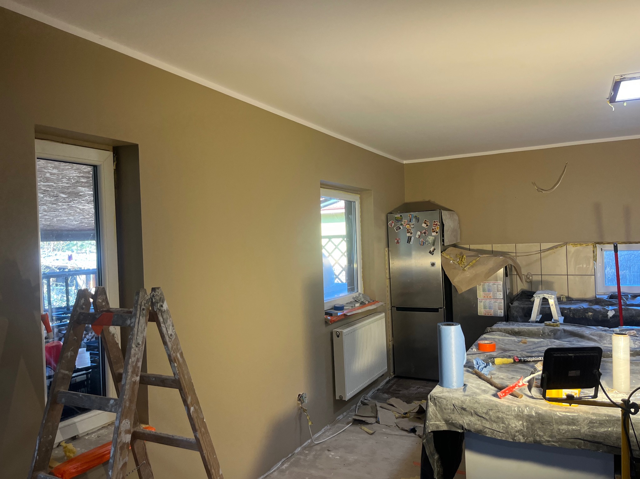 Kuchnia po malowaniu ścian na beżowo z białą listwą sufitową. Widoczne okno, drabina malarska i zabezpieczone meble. Ślady prac remontowych na podłodze.