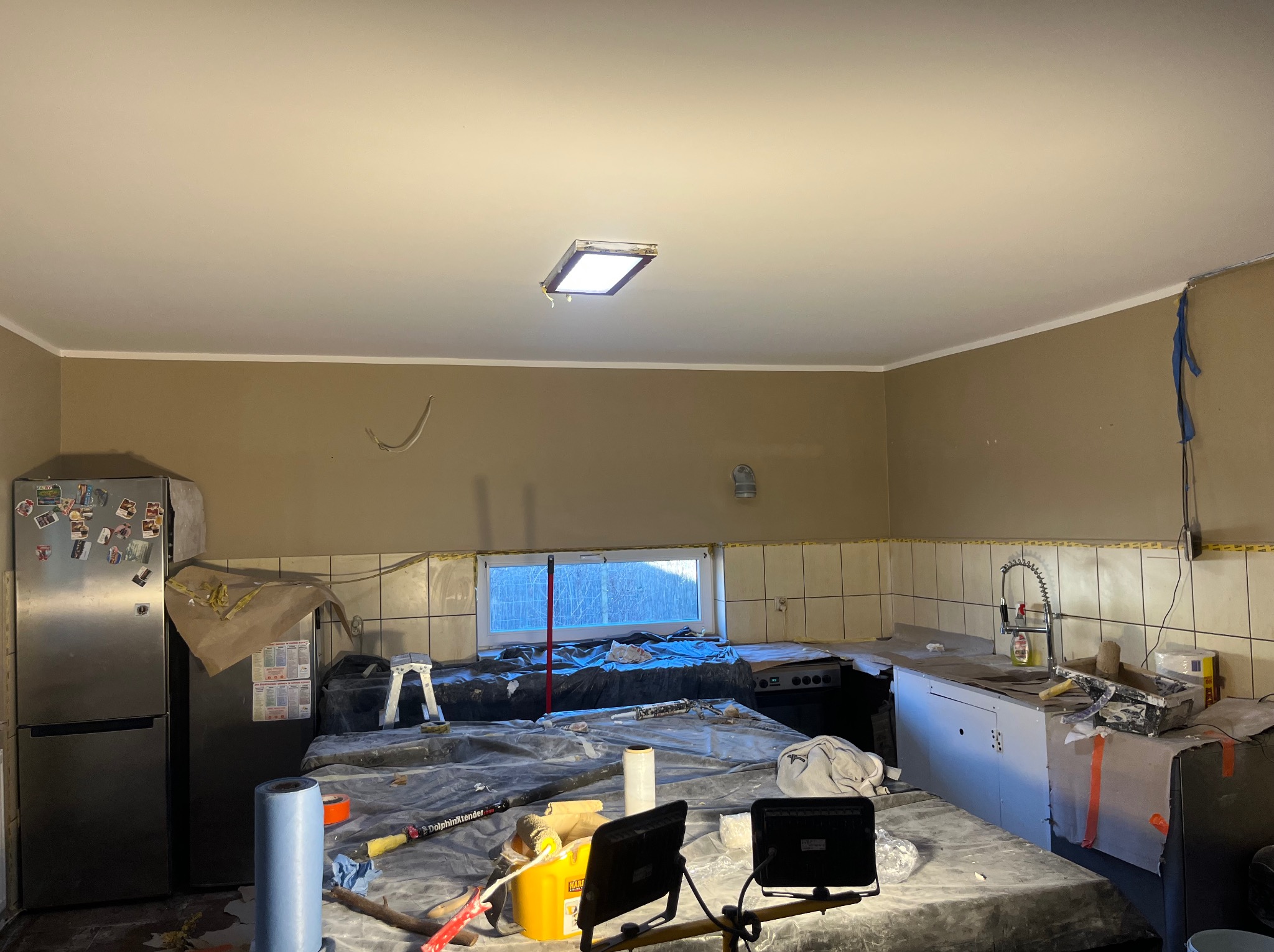 Przygotowania do malowania kuchni: zakryte meble folią, widoczne narzędzia malarskie i oświetlenie halogenowe. Całość w jasnym, roboczym świetle.