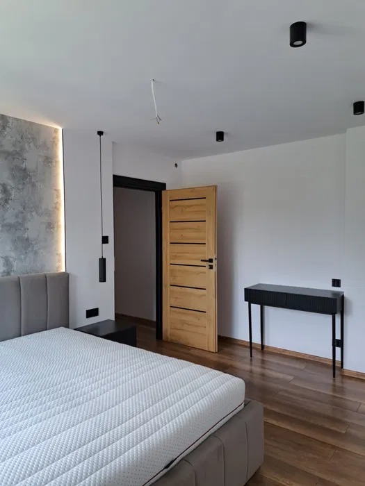 Nowoczesna sypialnia po remoncie: drewniana podłoga, łóżko z szarą ramą, ściana z efektem betonu, drewniane drzwi z czarną ościeżnicą i minimalistyczne oświetlenie.
